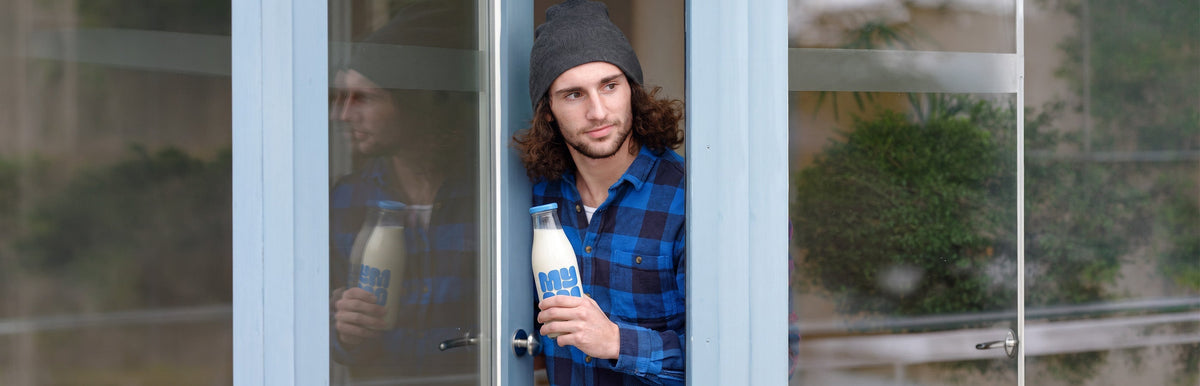 Man in a blue checked shirt standing in a doorway holding a glass bottle of MYOM oat milk.
