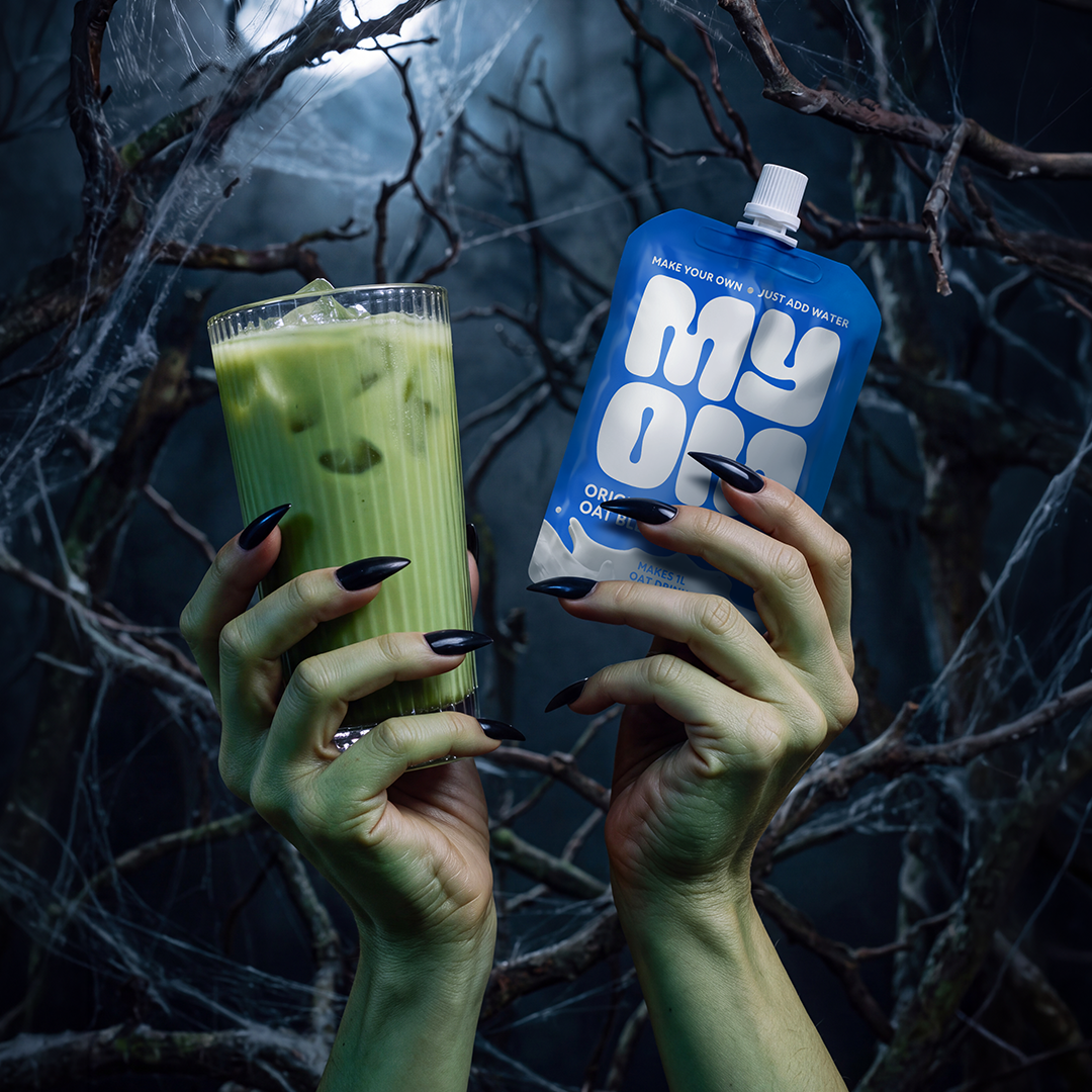 Green MYOM Monster Matcha over ice beside a MYOM Original Oat Drink Premix pouch, held by green hands with black nails.