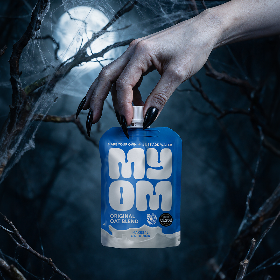 A hand with black nails holding a MYOM Original Oat Drink Premix pouch against a moonlit Halloween backdrop.
