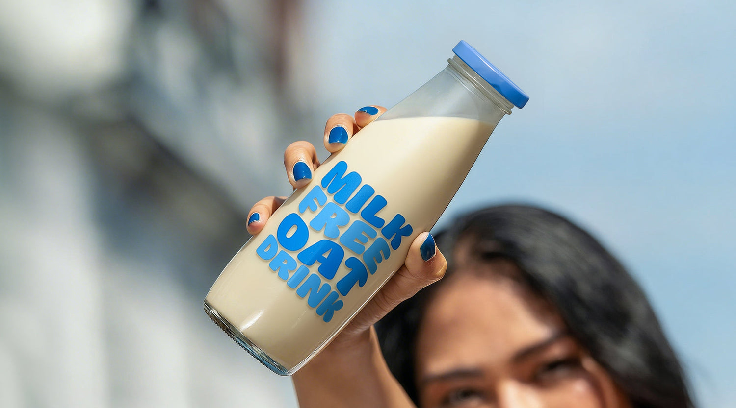 Woman raising a glass bottle of milk-free oat drink with bold blue lettering, outdoor lifestyle setting in daylight