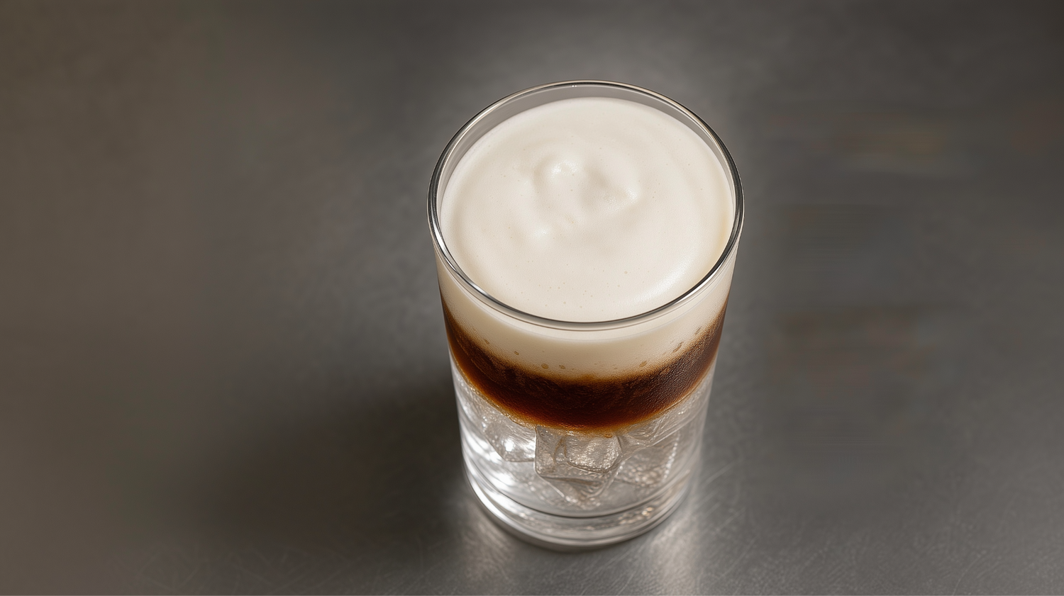 Top-down three-quarter view of a three-layer espresso tonic in a clean tumbler. Tonic-soaked ice at the base, a dark suspended espresso band, and a glossy MYOM Barista cold foam crown filling the top half.