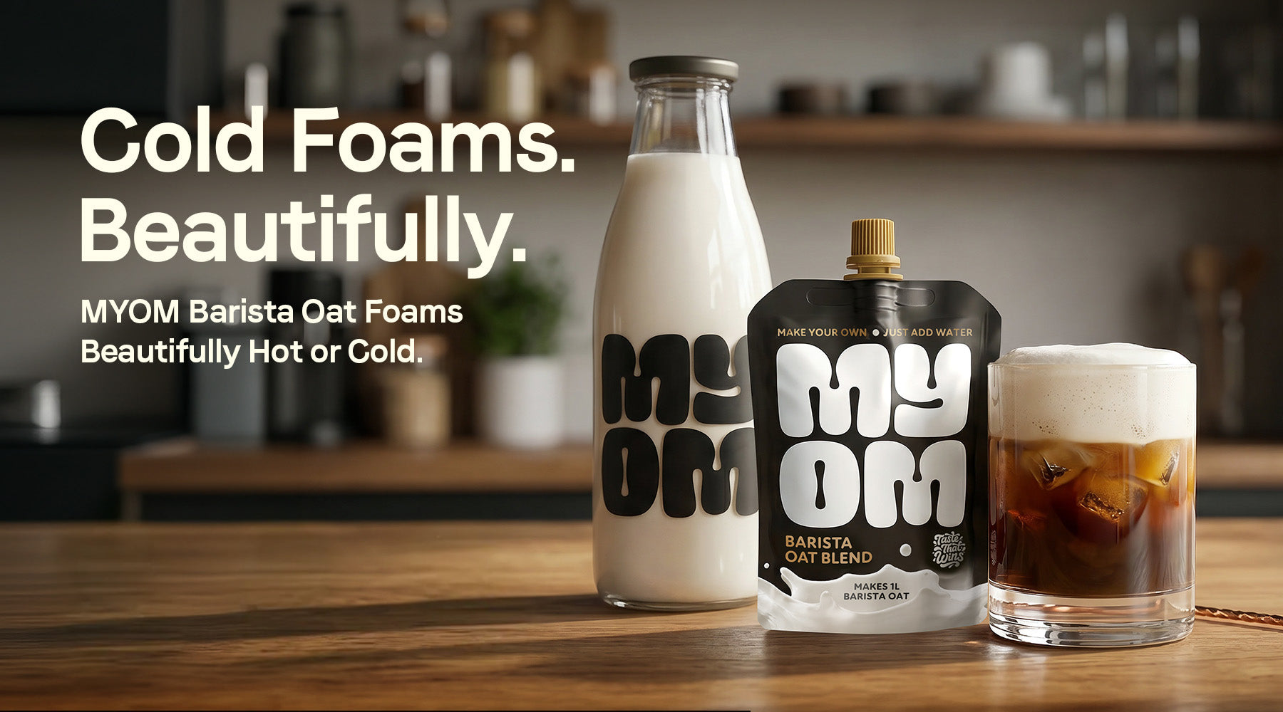 Cold Foams. Beautifully. MYOM Barista Oat pouch, glass bottle of fresh oat milk, and iced coffee with rich, creamy cold foam on a wooden kitchen counter.