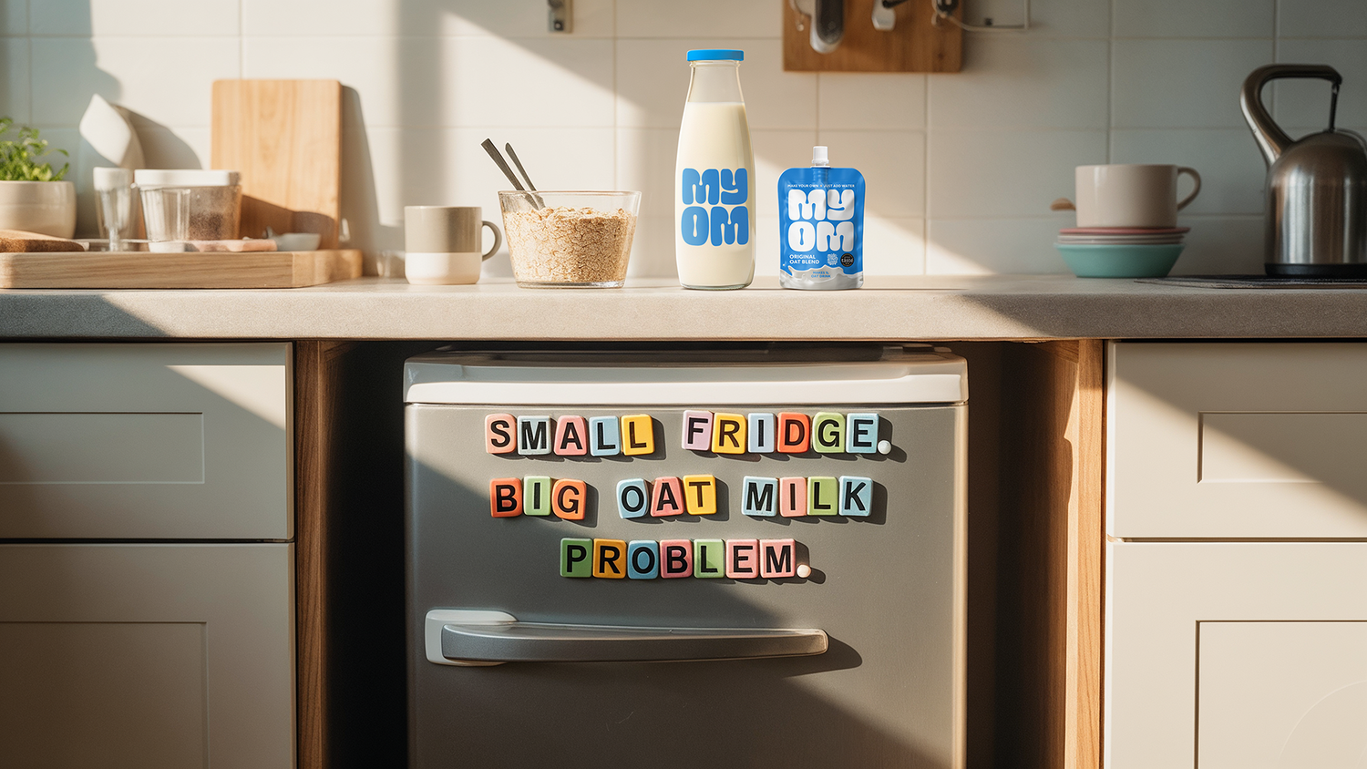 Small under-counter fridge in a shared kitchen with fridge magnets reading “Small fridge, big oat milk problem” and MYOM oat milk on the counter above.