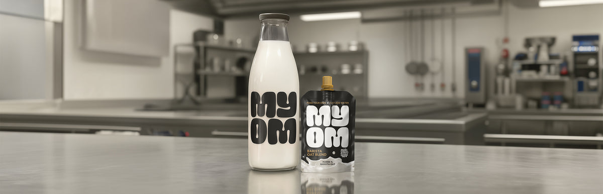 MYOM Barista oat milk premix pouch beside a filled 1L glass bottle on a stainless kitchen counter