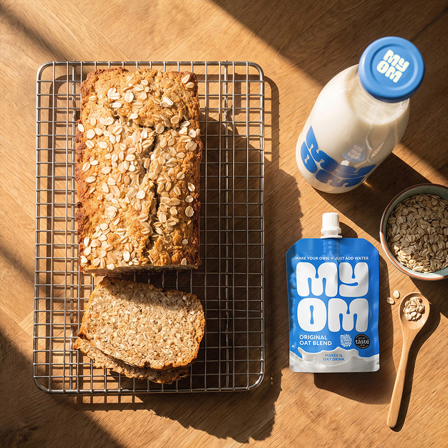 Overhead view of sliced porridge bread on a cooling rack with a MYOM Original Oat Drink Premix pouch, oat drink bottle and bowl of rolled oats.