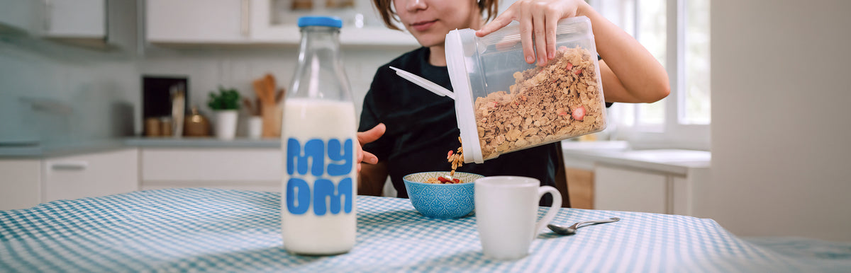 A lady pouring cereal into a bowl beside a glass bottle of MYOM oat milk at the breakfast table.