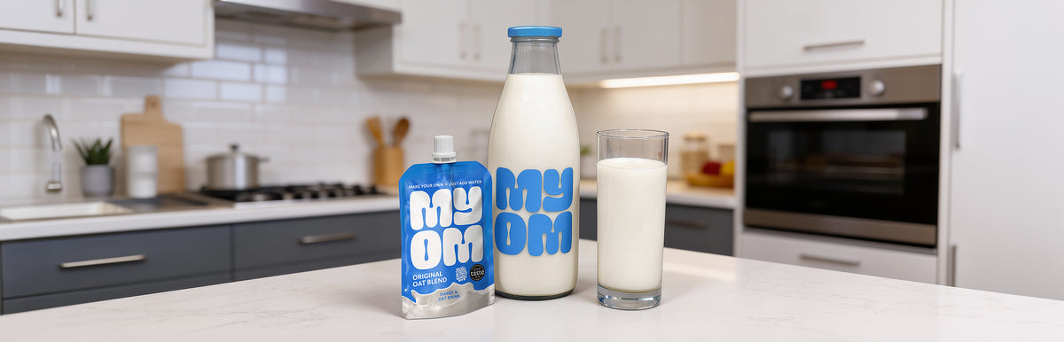 MYOM Original oat milk premix pouch beside a filled 1L glass bottle and a poured glass on a kitchen worktop