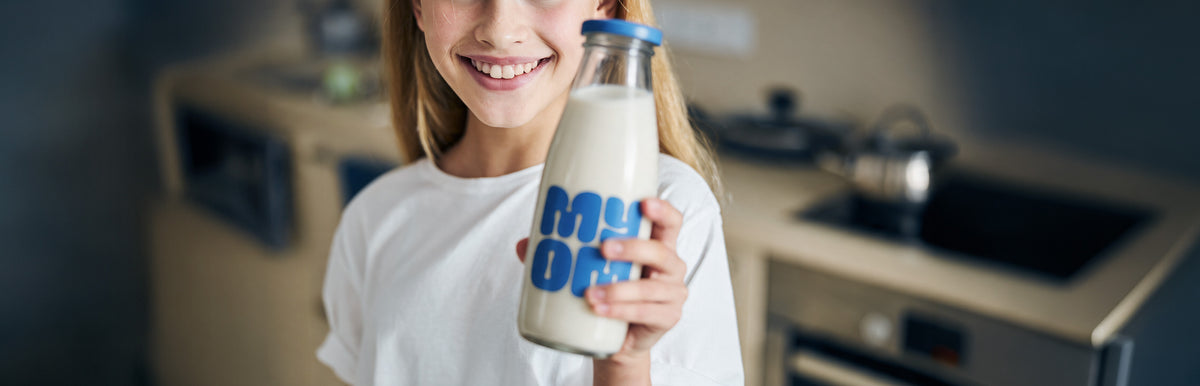 Smiling child holding a glass bottle of MYOM oat milk in a kitchen.